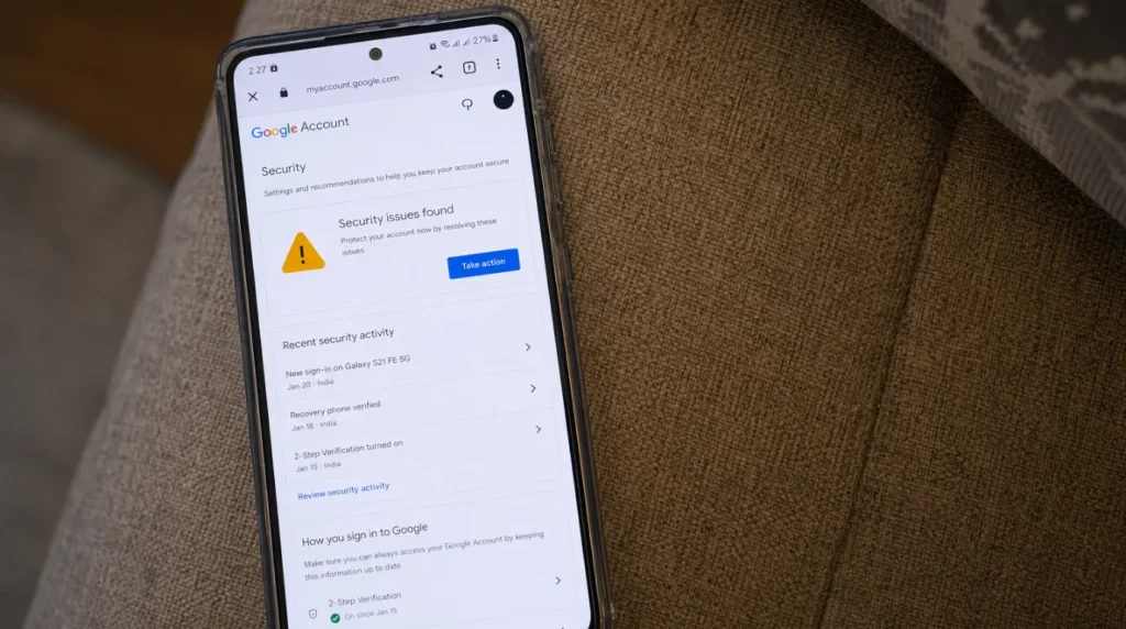A smartphone resting on a couch cushion showing a Google Account security alert page with a yellow warning triangle.